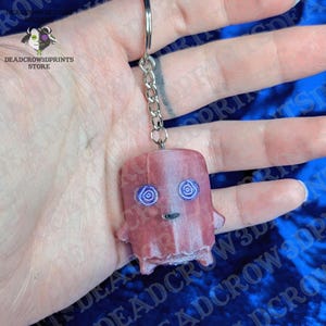 May include: A pink, semi-translucent keychain featuring a small, blocky character with two purple spiral eyes and a simple black mouth. The figure is attached to a silver-toned metal chain and a circular split ring for easy attachment to keys.
