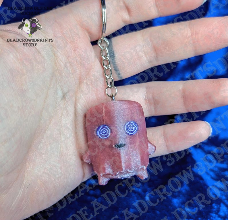 May include: A pink, semi-translucent keychain featuring a small, blocky character with two purple spiral eyes and a simple black mouth. The figure is attached to a silver-toned metal chain and a circular split ring for easy attachment to keys.