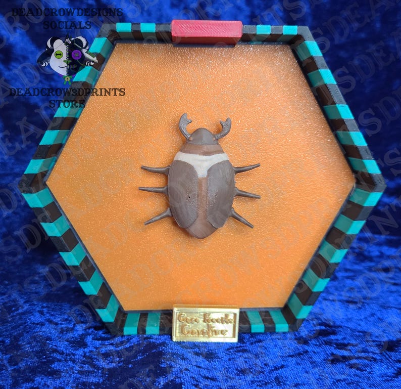 a 3d printed brown and tan coco beetle from the movie coraline in a green, black and orange button box with a gold plate that says "coco beetle Coraline"
