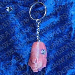 May include: A keychain featuring a metallic split ring and a short chain attached to a pink, textured, elongated object with a single, small, blue spiral-shaped eye detail on its surface. The object has a smooth, slightly iridescent finish.