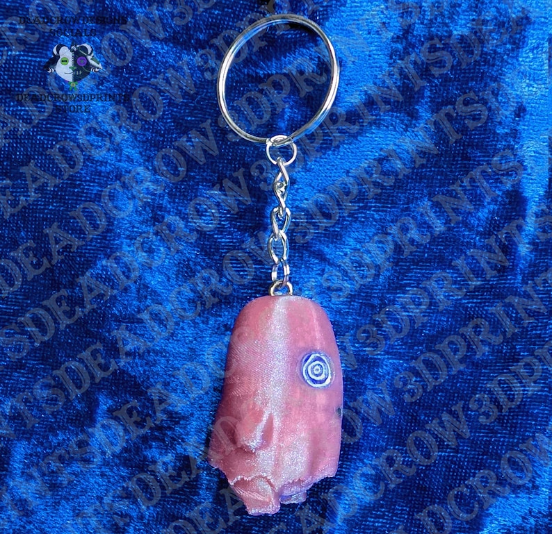 May include: A keychain featuring a metallic split ring and a short chain attached to a pink, textured, elongated object with a single, small, blue spiral-shaped eye detail on its surface. The object has a smooth, slightly iridescent finish.