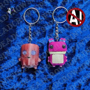 May include: Two keychains featuring stylized figures. The left figure is a reddish-brown ghost with blue spiral eyes. The right figure is a bright pink creature with large black eyes, blue ears, and a white rectangular chest panel, both on metal rings.