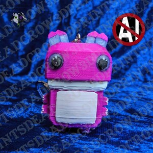 May include: A 3D printed keychain featuring a blocky, bright pink creature with large black eyes, light blue ears, and a white rectangular patch on its chest. The figure has a textured surface and a metal ring attached to the top for hanging.