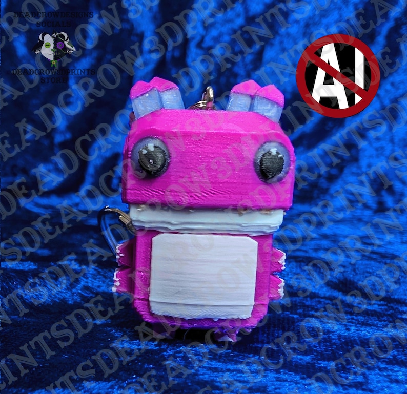 May include: A 3D printed keychain featuring a blocky, bright pink creature with large black eyes, light blue ears, and a white rectangular patch on its chest. The figure has a textured surface and a metal ring attached to the top for hanging.