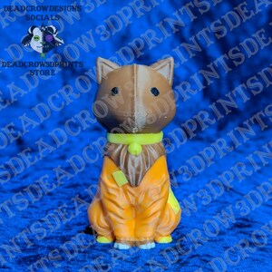 May include: A 3D printed figurine of a brown dog with pointed ears and black eyes. The dog wears a bright yellow collar with a small bell and an orange jumpsuit with a yellow patch on the chest. The figure has a visible vertical seam line.