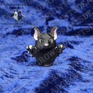 May include: A black cat-shaped 3D-printed item with white and pink accents. The cat has its paws outstretched. The item is a decorative piece or a functional holder. The text "DEADCROWDESIGNS" and "STORE" are visible.
