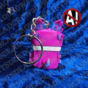 May include: A 3D-printed keychain featuring a bright pink blocky character with a white horizontal stripe across the middle. The figure has small purple-tipped ears and a purple-tipped tail, attached to a silver metal split ring keychain.