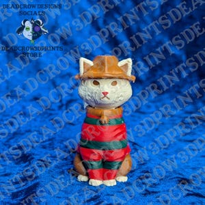 May include: A 3D printed figurine of a white cat wearing a brown fedora hat and a red and green horizontally striped sweater. The cat has pointed ears, a small red nose, and a neutral expression. The figure is rendered in a solid, matte finish.