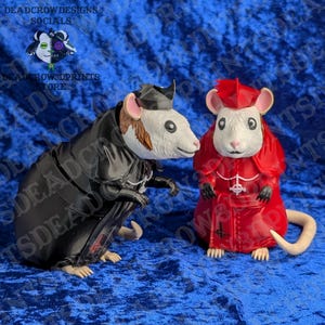 May include: Two rat figurines, one wearing a black robe and hat, the other wearing a red robe and hat. Both have white fur, pink ears, and black eyes. The red-robed rat features a white cross symbol on the chest and a tail extending to the right.
