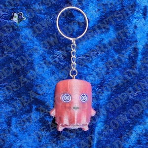 May include: A keychain featuring a cylindrical, reddish-pink character figure with two purple spiral eyes and a small mouth. The figure is attached to a silver-toned metal split ring by a short chain. The surface of the figure has a textured finish.