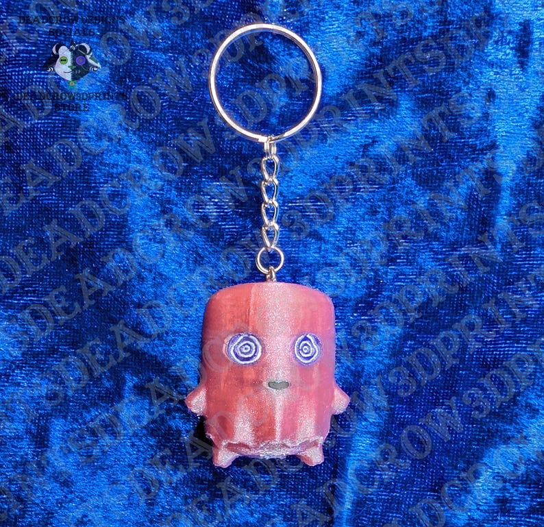 May include: A keychain featuring a cylindrical, reddish-pink character figure with two purple spiral eyes and a small mouth. The figure is attached to a silver-toned metal split ring by a short chain. The surface of the figure has a textured finish.