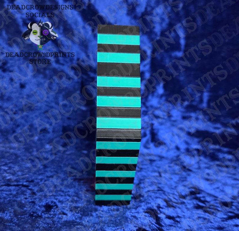 Puede incluir: Un objeto rectangular vertical con rayas horizontales alternas negras y turquesas. El objeto se encuentra sobre un fondo azul. El texto "DEADCROWDESIGNS SOCIALS" y "DEADCROW3DPRINTS STORE" son visibles.
