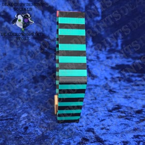 Puede incluir: Un objeto rectangular y alto con rayas negras y turquesas alternas. El objeto est&aacute; hecho de capas apiladas, con un acento rojo en la parte superior. El fondo es una tela de terciopelo azul oscuro. El texto a la izquierda dice "DEADCROW DESIGNS SOCIALS" y "DEADCROW3DPRINTS STORE".