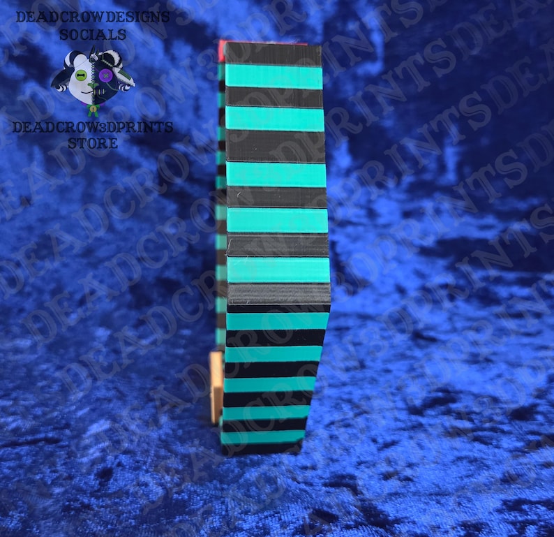 Puede incluir: Un objeto rectangular y alto con rayas negras y turquesas alternas. El objeto est&aacute; hecho de capas apiladas, con un acento rojo en la parte superior. El fondo es una tela de terciopelo azul oscuro. El texto a la izquierda dice "DEADCROW DESIGNS SOCIALS" y "DEADCROW3DPRINTS STORE".