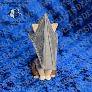 May include: A 3D printed figurine of a creature wearing a large, grey, triangular pyramid-shaped headpiece. The figure has light-colored paws and brown lower body sections, with the pyramid structure featuring visible horizontal layer lines.
