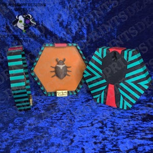 Puede incluir: Colecci&oacute;n de cajas hexagonales decorativas y una pulsera. Las cajas presentan un escarabajo sobre un fondo naranja, enmarcadas en rayas negras y turquesas. La pulsera tiene un dise&ntilde;o de rayas similar. El texto "Deadrow Designs" es visible.