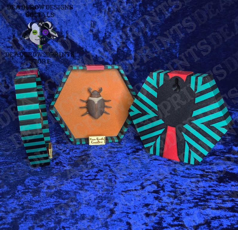 Puede incluir: Colecci&oacute;n de cajas hexagonales decorativas y una pulsera. Las cajas presentan un escarabajo sobre un fondo naranja, enmarcadas en rayas negras y turquesas. La pulsera tiene un dise&ntilde;o de rayas similar. El texto "Deadrow Designs" es visible.