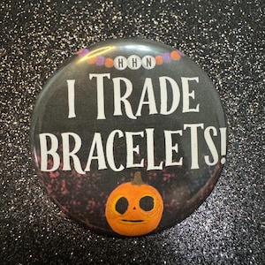 May include: A round button with the text "I TRADE BRACELETS!" in white, with a cartoon jack-o'-lantern at the bottom. The button has a black background with a glittery effect and the letters "HHN" at the top.