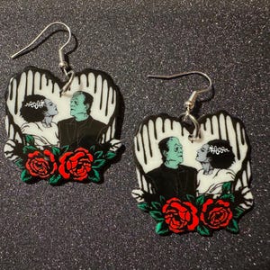 May include: A pair of black and white heart-shaped earrings with a graphic print of Frankenstein and his bride. The earrings feature red roses and silver hooks.