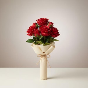 May include: A bouquet of red roses with green leaves, wrapped in cream-colored paper and tied with twine. The roses are in full bloom, showcasing their vibrant color. The arrangement is presented on a white surface against a neutral background.
