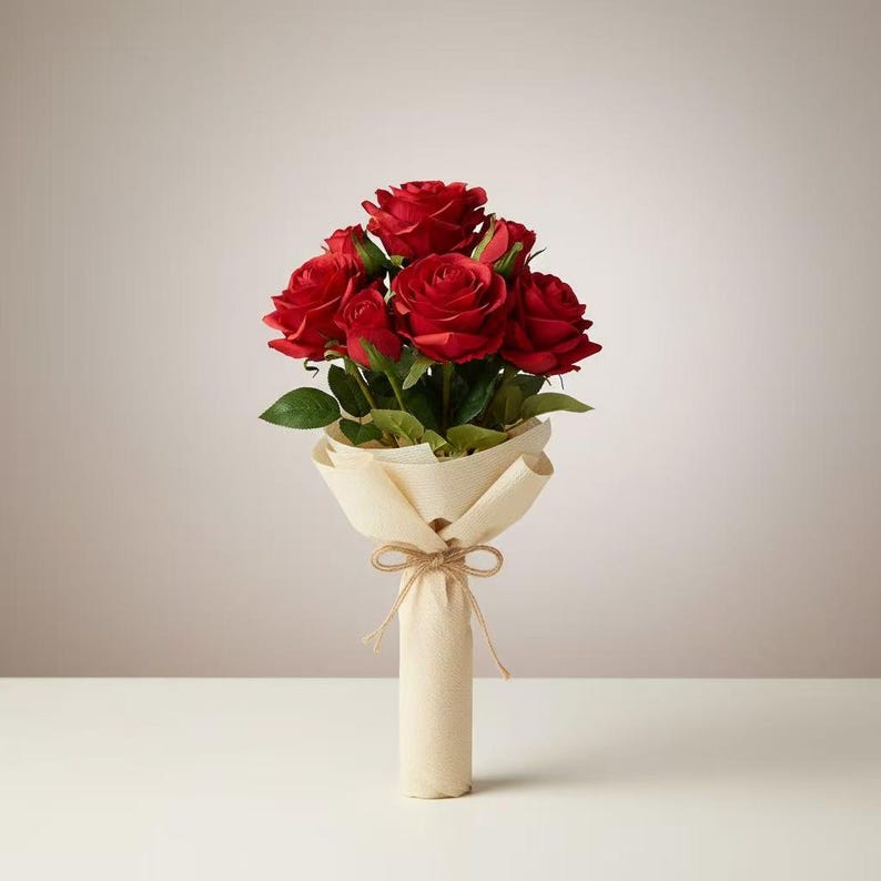 May include: A bouquet of red roses with green leaves, wrapped in cream-colored paper and tied with twine. The roses are in full bloom, showcasing their vibrant color. The arrangement is presented on a white surface against a neutral background.