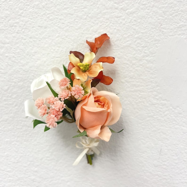 Puede incluir: Un prendido floral con una rosa color melocot&oacute;n como punto focal, complementada con flores rosas y blancas m&aacute;s peque&ntilde;as, y acentuada con follaje naranja y verde. Una cinta blanca asegura los tallos.
