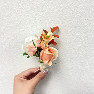 Puede incluir: Un prendedor floral con una rosa melocot&oacute;n como punto central, complementada con flores blancas, naranjas y rosas. Hojas verdes y acentos de follaje naranja completan el arreglo. Se adjunta una peque&ntilde;a cinta blanca.
