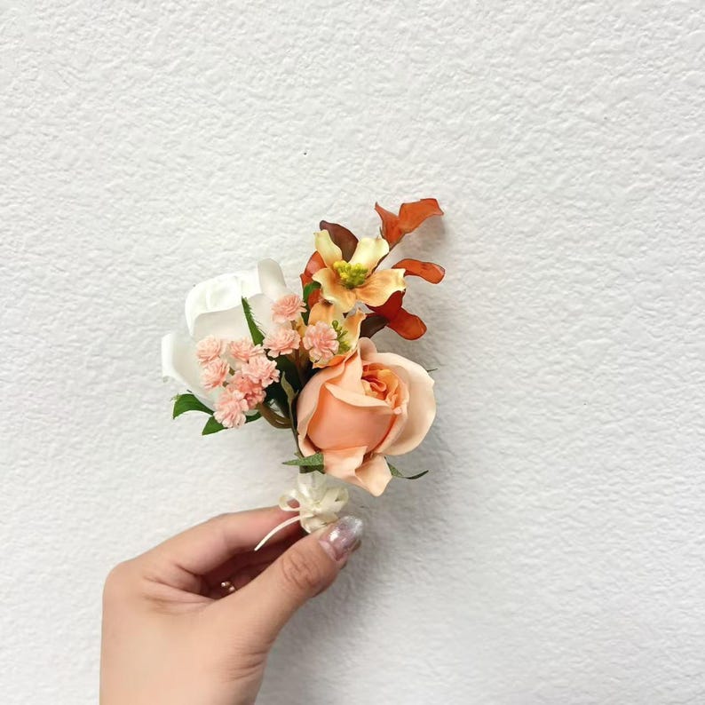 Puede incluir: Un prendedor floral con una rosa melocot&oacute;n como punto central, complementada con flores blancas, naranjas y rosas. Hojas verdes y acentos de follaje naranja completan el arreglo. Se adjunta una peque&ntilde;a cinta blanca.