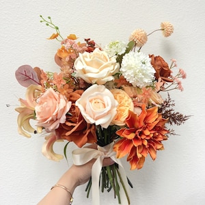 Puede incluir: Un vibrante ramo de flores artificiales en tonos melocot&oacute;n, naranja y crema. El arreglo incluye rosas, dalias y otras flores, atado con una cinta color crema. El ramo es sostenido en la mano de una persona.