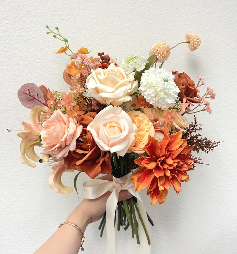 Puede incluir: Un vibrante ramo de flores artificiales en tonos melocot&oacute;n, naranja y crema. El arreglo incluye rosas, dalias y otras flores, atado con una cinta color crema. El ramo es sostenido en la mano de una persona.
