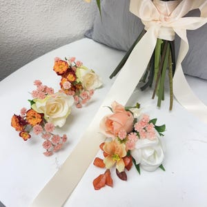 Puede incluir: Arreglo floral con un ramo con una cinta color crema y varias piezas florales m&aacute;s peque&ntilde;as. Los arreglos m&aacute;s peque&ntilde;os incluyen rosas en tonos melocot&oacute;n, naranja y blanco, con toques de rosa y verde.