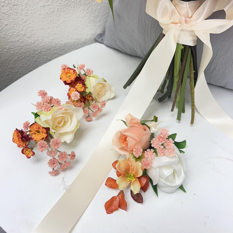 Puede incluir: Arreglo floral con un ramo con una cinta color crema y varias piezas florales m&aacute;s peque&ntilde;as. Los arreglos m&aacute;s peque&ntilde;os incluyen rosas en tonos melocot&oacute;n, naranja y blanco, con toques de rosa y verde.
