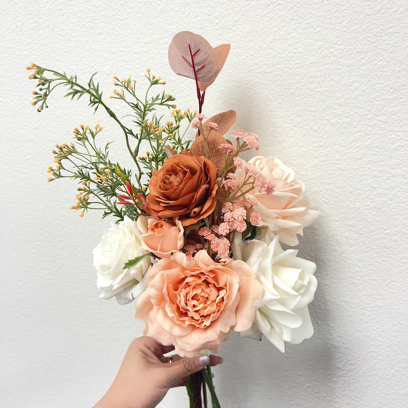 Puede incluir: Un ramo de flores artificiales con rosas melocot&oacute;n, blancas y naranja quemado, acentuado con vegetaci&oacute;n y peque&ntilde;as flores rosas. El arreglo presenta una variedad de texturas y colores, creando un aspecto natural y elegante.