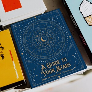 May include: A blue book with a gold zodiac wheel and constellations. The title is "A Guide to Your Stars" with the subtitle "2024 Personalized Horoscopes".