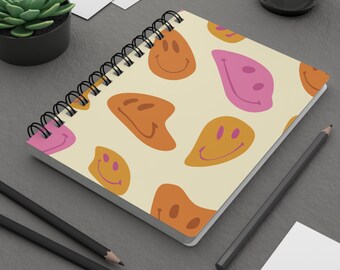 May include: Spiral-bound notebook featuring a cream-colored cover adorned with a repeating pattern of distorted smiley faces in shades of orange and pink. The notebook is open, revealing blank pages, and rests on a gray surface with pencils and a small plant.