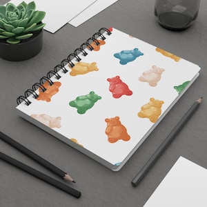 Op de afbeelding: Een spiraalgebonden notitieboek met een herhalend patroon van kleurrijke gummybeer illustraties. De beren zijn in verschillende kleuren, waaronder rood, groen, geel, oranje en blauw, tegen een witte achtergrond. Het notitieboek heeft een zwarte spiraalbinding.