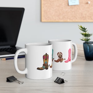 Puede incluir: Dos tazas de cerámica blanca con un diseño de botas de vaquero coloridas. Las botas están decoradas con motivos florales y tienen espuelas. Las tazas están sobre una mesa de madera.