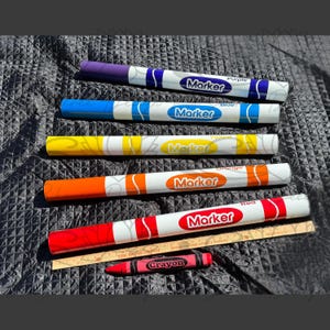 May include: An assortment of colorful markers and a crayon are displayed. The markers are labeled with the colors purple, blue, yellow, orange, and red. A wooden ruler is also present. The crayon is red and labeled "Crayon."