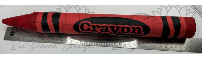 A large red crayon with black stripes and the word 'Crayon' printed in black letters on the side. The crayon is lying on a silver ruler showing the 15” length.