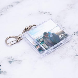 May include: A square, clear plastic keychain case featuring a miniature CD-style insert. The insert displays a photograph of two people standing outdoors near a Ferris wheel. The keychain includes a silver-toned metal ring and a lobster clasp.