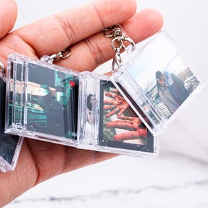 May include: A clear plastic keychain featuring miniature CD jewel cases connected together. Each case displays a different printed photograph, including a person in a dark setting, a close-up of hands, and a couple standing outdoors together.