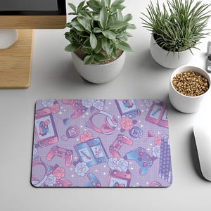 May include: A rectangular mouse pad featuring a repeating pattern of gaming consoles, controllers, cat-ear headphones, and floral motifs in shades of pastel pink, blue, and purple against a light grid background with small white star accents.