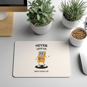May include: A rectangular off-white mouse pad featuring a cartoon illustration of a yellow handheld video game console with a face, arms, and legs. The text above reads NEVER GIVE UP, and the text below reads ONLY LEVEL UP! in black font.