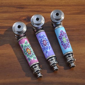 May include: Three metal smoking pipes with colourful floral designs. The pipes are silver with a pink, purple, and blue floral pattern.
