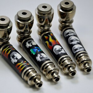 May include: Four metal smoking pipes with different skull and flame designs. The pipes are silver with black and colorful accents.