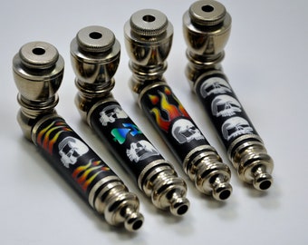 May include: Four metal smoking pipes with different skull and flame designs. The pipes are silver with black and colourful accents.