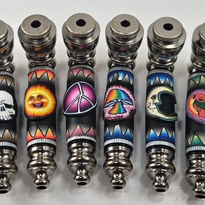 May include: Seven metal smoking pipes with colorful, decorative designs. The designs include a skull, a smiling sun, a peace sign, a mushroom, a crescent moon, and a dancing bear.