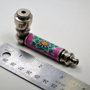 May include: A silver metal smoking pipe with a pink and green floral design. The pipe is approximately 10 cm long and has a bowl at one end and a mouthpiece at the other.