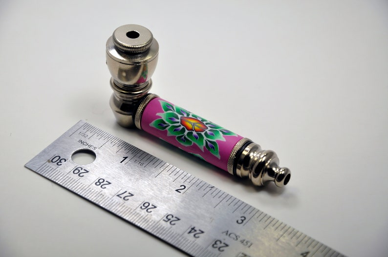 May include: A silver metal smoking pipe with a pink and green floral design. The pipe is approximately 10 cm long and has a bowl at one end and a mouthpiece at the other.