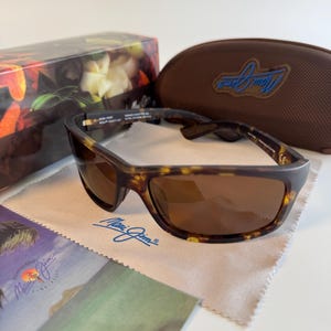 May include: Tortoiseshell-patterned sunglasses with brown lenses, featuring the Maui Jim logo on the temple. Includes a brown hard case with a blue logo, a branded microfiber cleaning cloth, and a floral-patterned box with product information.